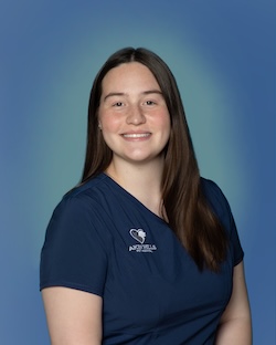 A woman with long brown hair wearing navy blue medical scrubs stands in front of a blue gradient background, smiling at the camera.