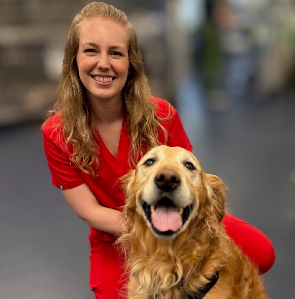 A woman in red scrubs smiles while kneeling next to a happy golden retriever indoors.